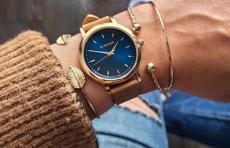 Gold-toned wristwatch with blue face and brown leather band, worn on a wrist with gold bracelets.