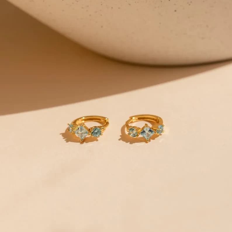 A pair of gold-colored hoop earrings, each adorned with three light blue gemstones.