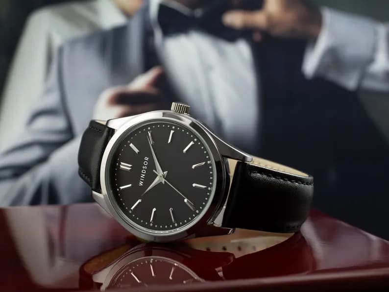 A silver watch with a black leather band and a black face, featuring silver hands and Roman numeral hour markers. The word 'WINDSOR' is printed on the face.