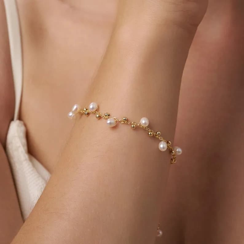 Gold chain bracelet with small white pearls hanging from it, worn on a wrist.
