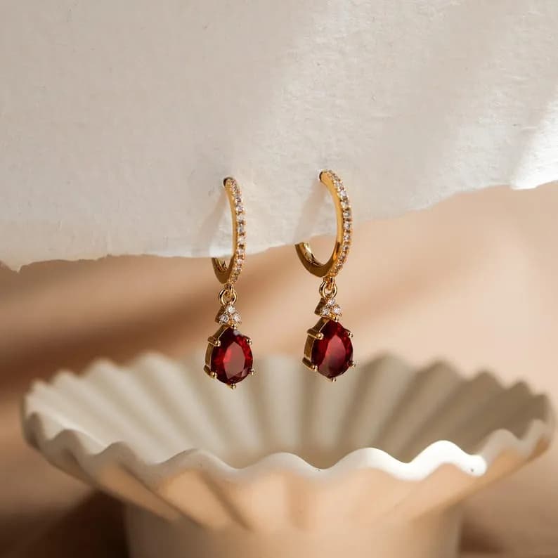 Gold hoop earrings with deep red gemstones and clear stones, displayed on a white dish.