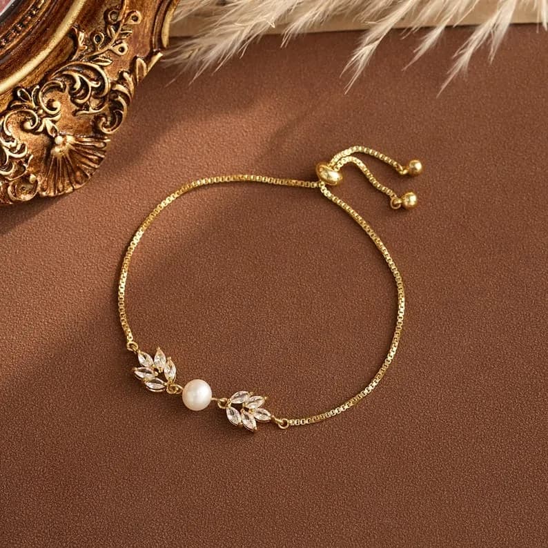 Gold-toned bracelet with a delicate chain and a sliding clasp featuring a central pearl and leaf-shaped accents adorned with clear crystals.
