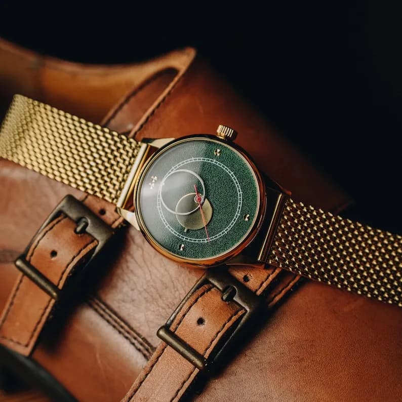 Gold-toned wristwatch with a green face and gold mesh band on a brown leather shoe.