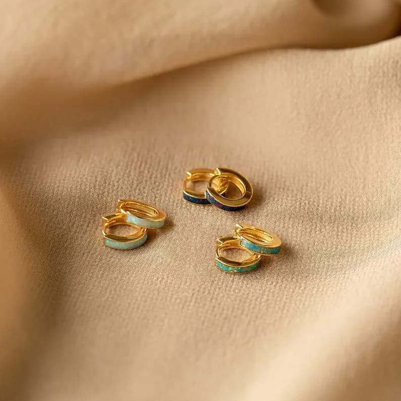 Four pairs of gold hoop earrings with different colored opal inlays arranged on a beige fabric background.