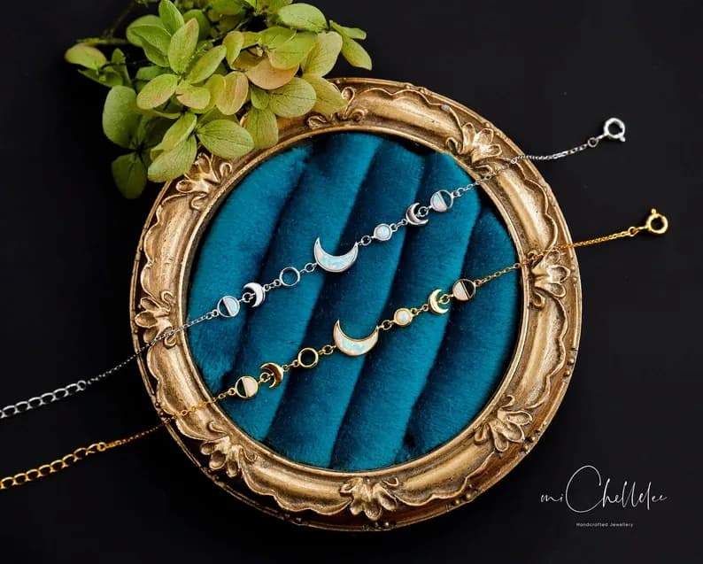 Delicate bracelets featuring moon phase designs with light blue and white iridescent finish on a teal velvet surface.