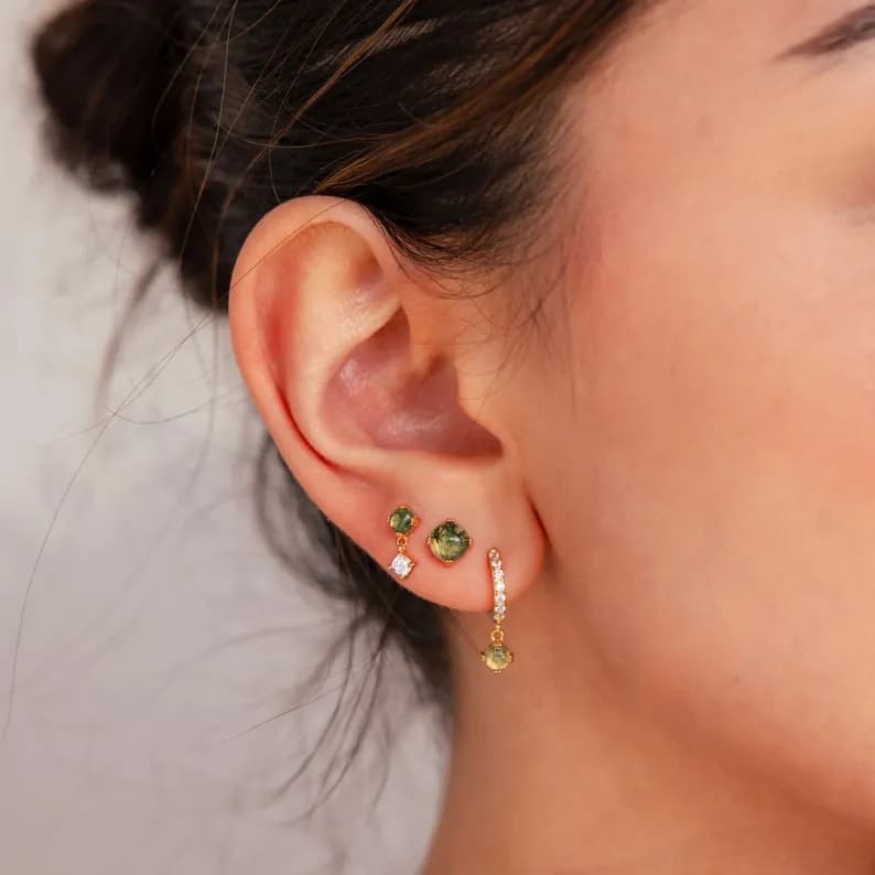 Close-up of a person's ear with three gold earrings featuring green and clear gemstones.