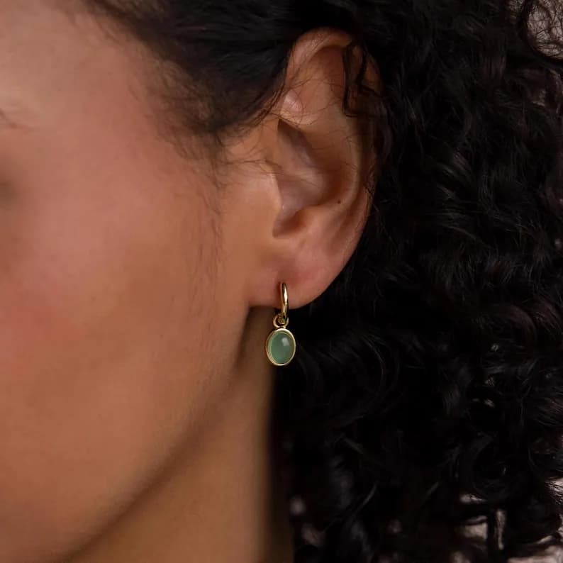 A pair of gold hoop earrings with a small oval-shaped green gemstone pendant hanging from each hoop.