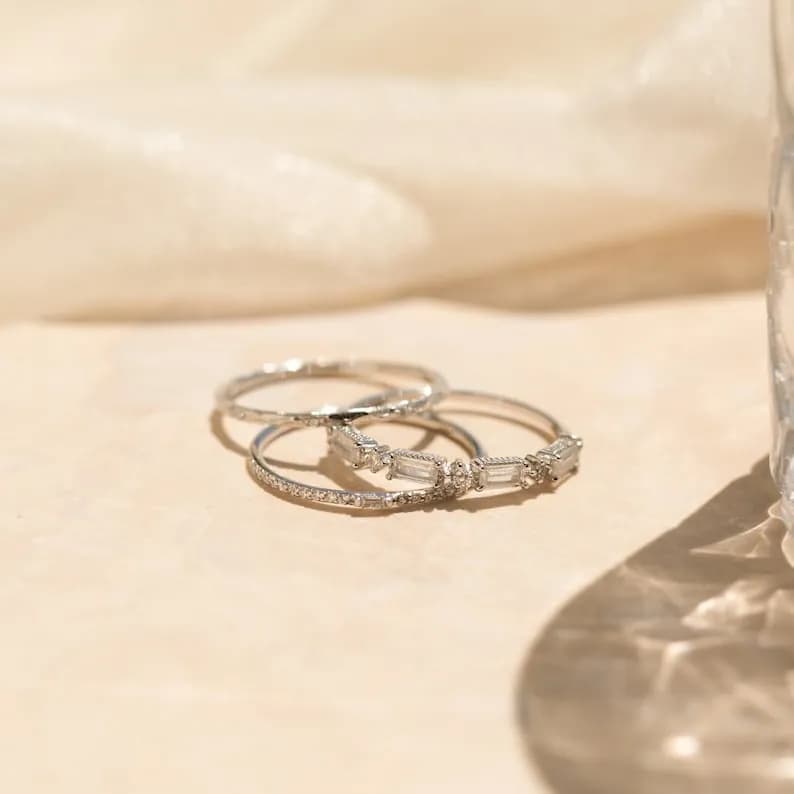 Three silver rings with small diamonds. The top ring has a row of five baguette-shaped diamonds. The bottom two rings are thin and have a delicate diamond pattern.