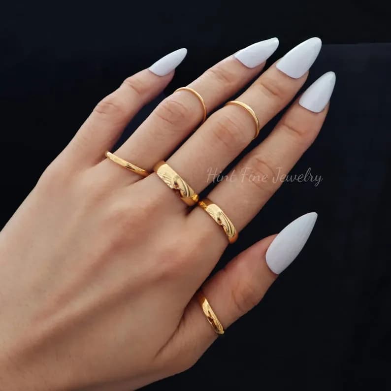 A hand wearing five gold rings, including a textured band, on three fingers with white nail polish.